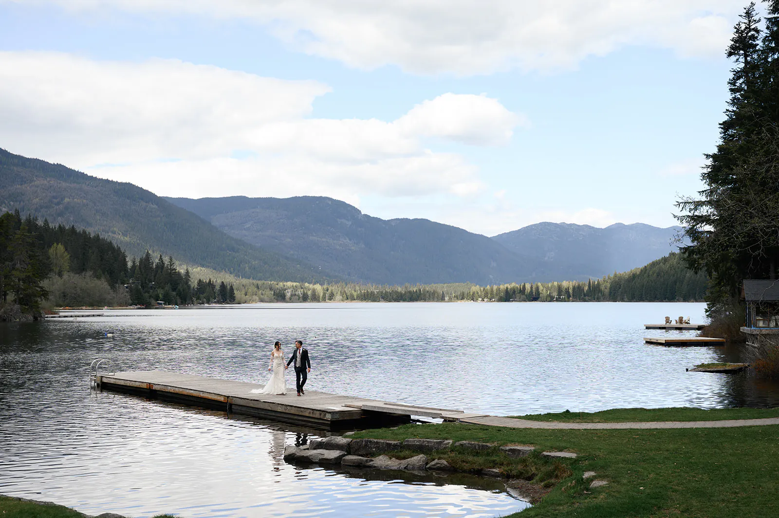 A bride and groom walk hand in hand on a wooden dock extending into a calm lake, surrounded by forested mountains under a partly cloudy sky.