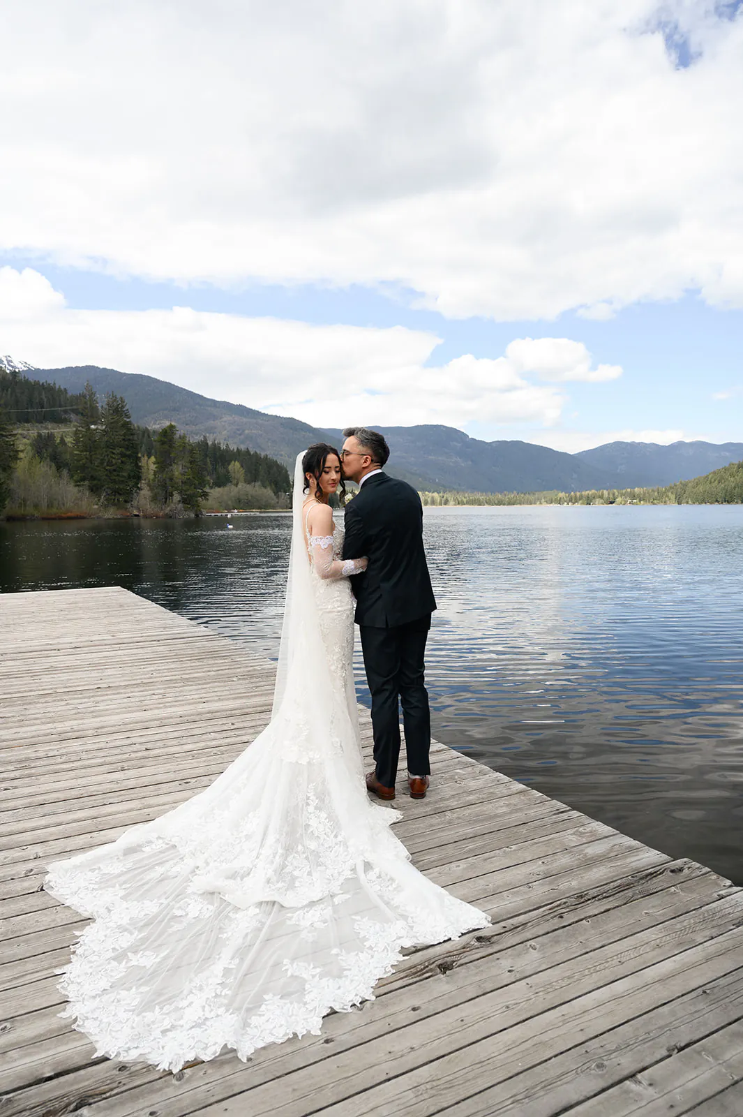 A bride and groom stand on a wooden dock by a lake, with mountains and trees in the background; the groom kisses the bride on the forehead.