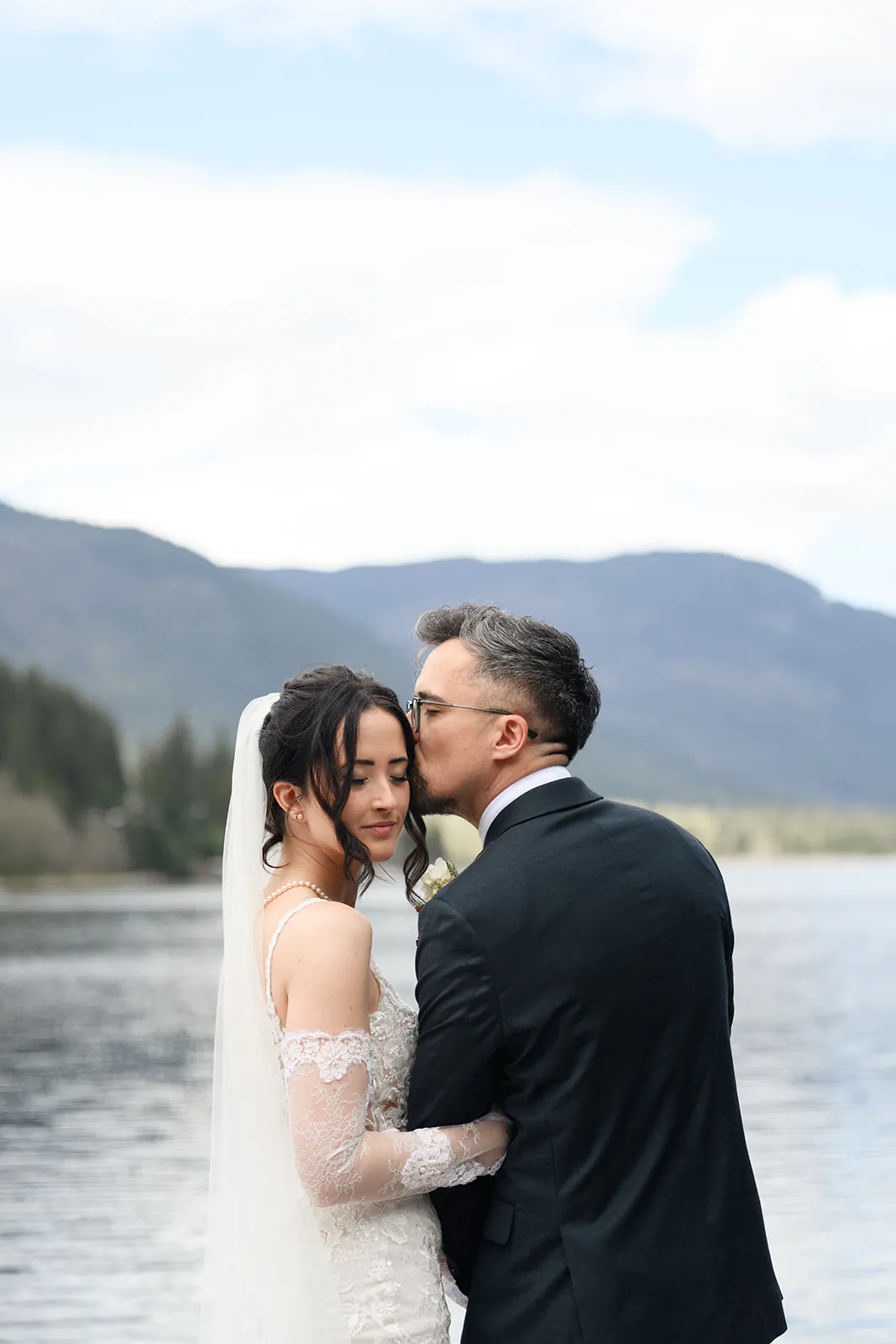 A bride and groom embrace by a lake, with mountains and trees in the background. The groom kisses the bride’s forehead while she closes her eyes.