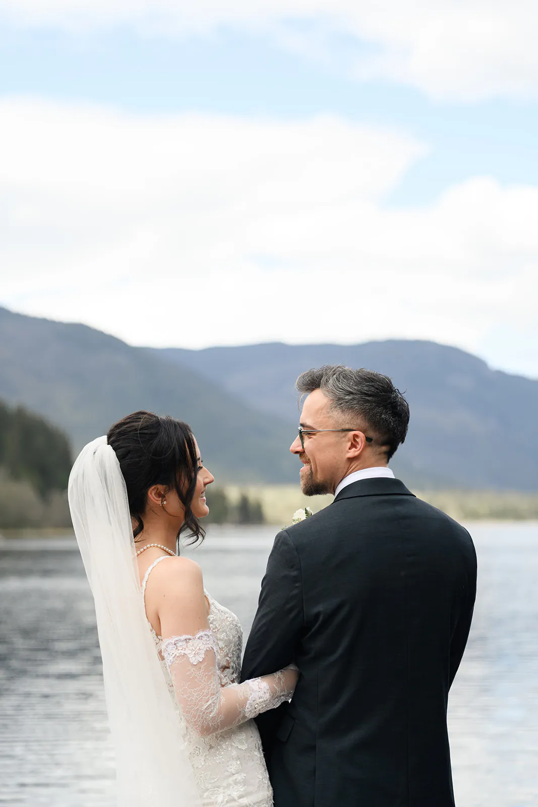 A bride and groom stand by a lake, facing each other and smiling, with mountains and cloudy sky in the background.