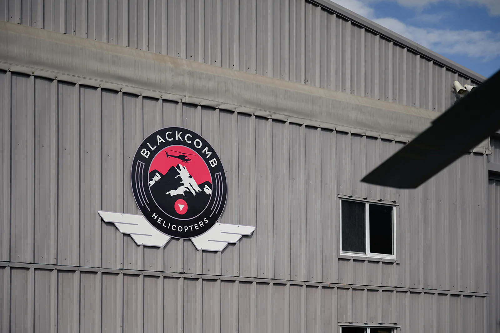 A metal building features a round "Blackcomb Helicopters" logo with a mountain and helicopter graphic; two windows are visible, and a helicopter blade is partially shown on the right.