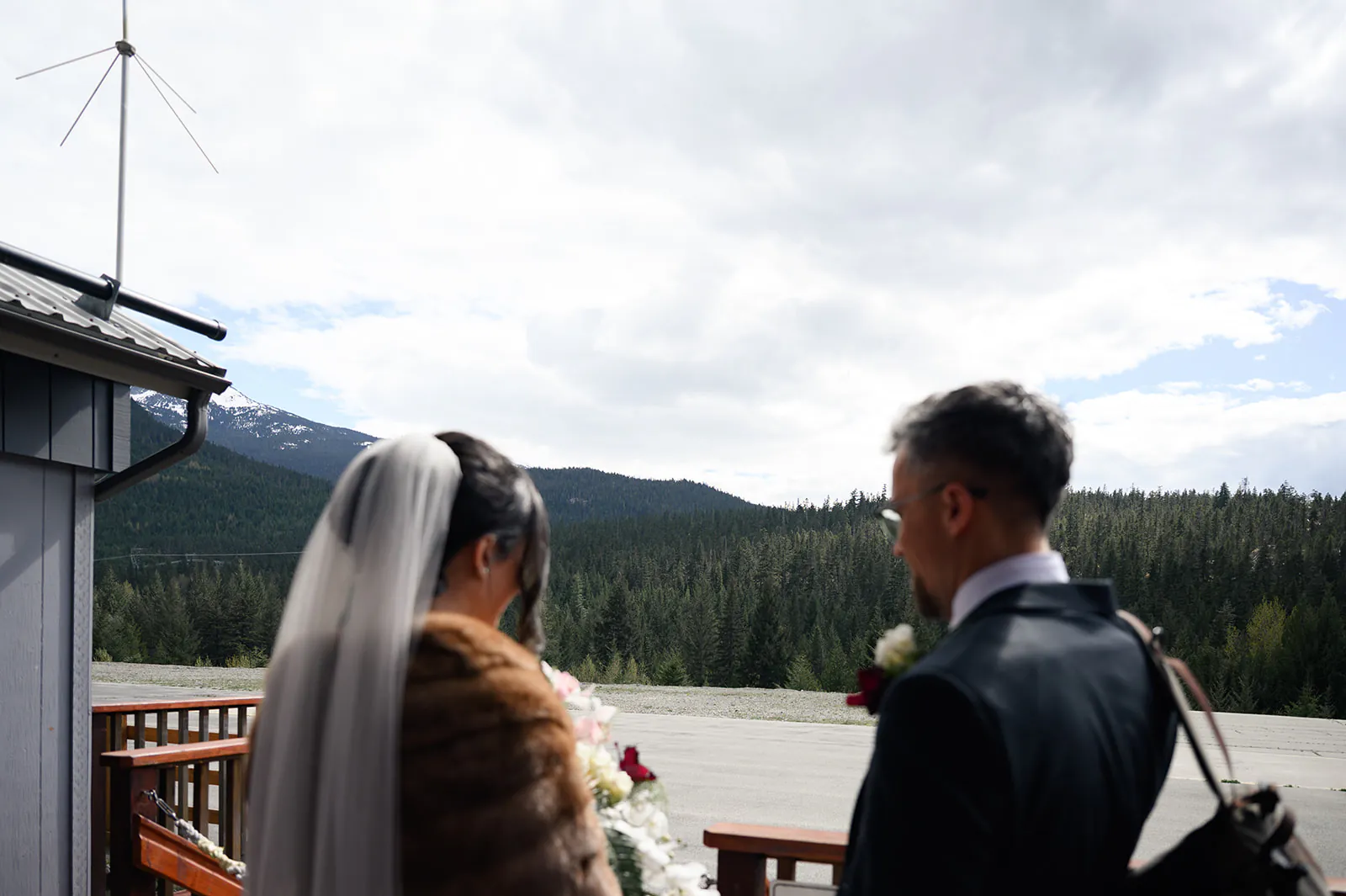 A bride and groom stand on a wooden deck overlooking a forest and mountains under a partly cloudy sky.