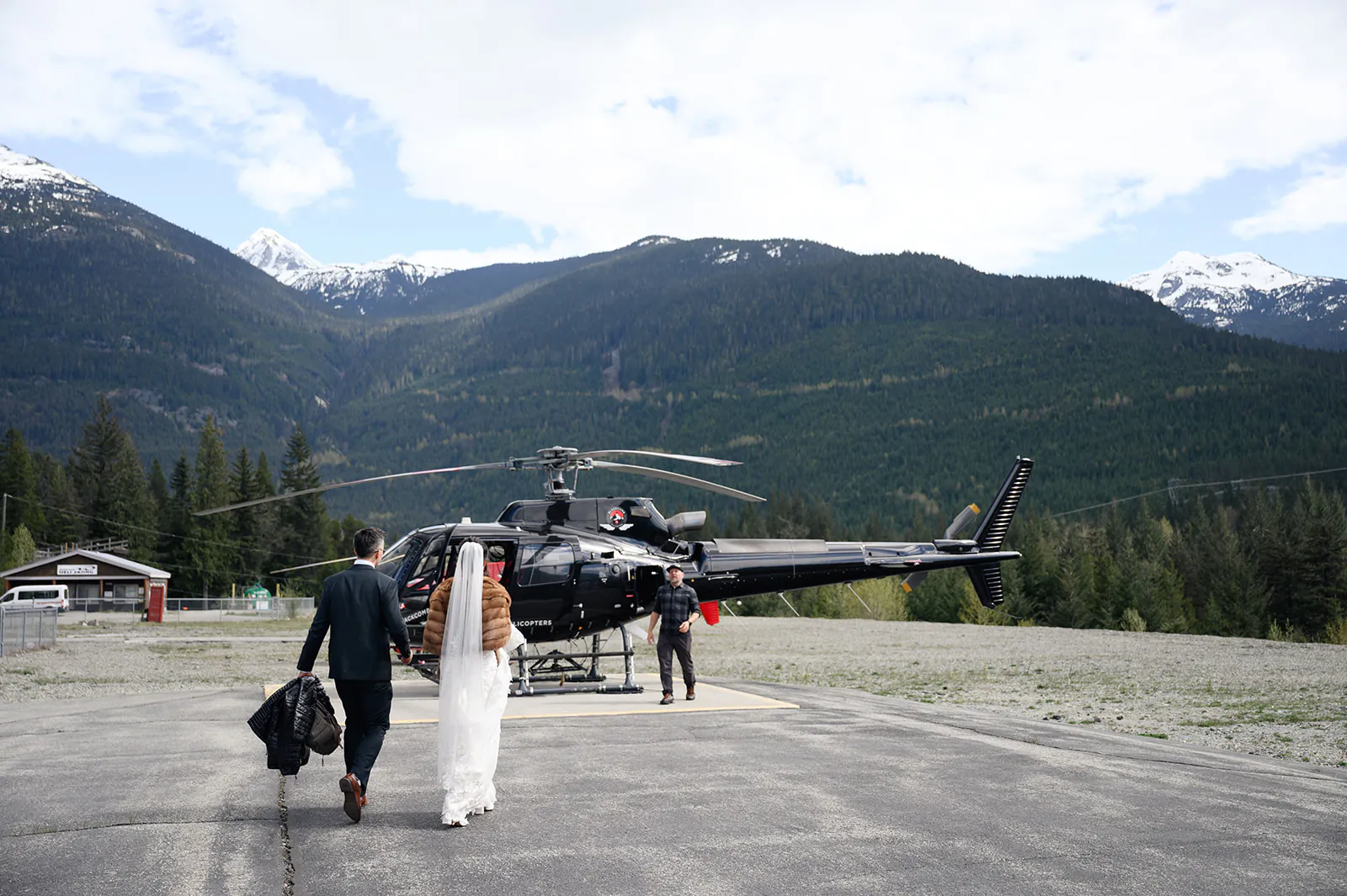 A bride and groom walk toward a black helicopter on a landing pad in a mountainous area, carrying bags, with another person near the helicopter.