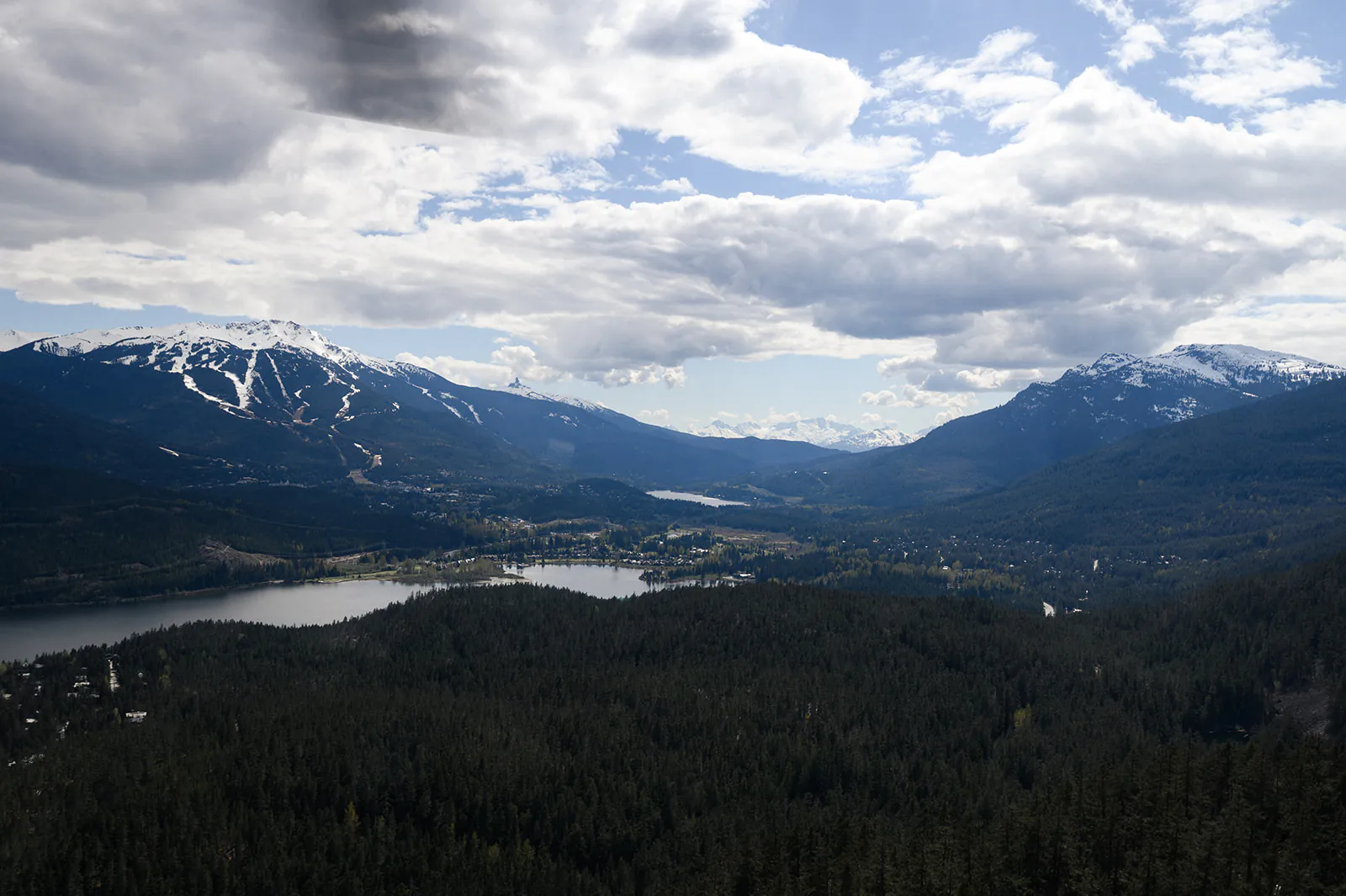 A scenic view of a forested valley with a lake, surrounded by snow-capped mountains under a partly cloudy sky.
