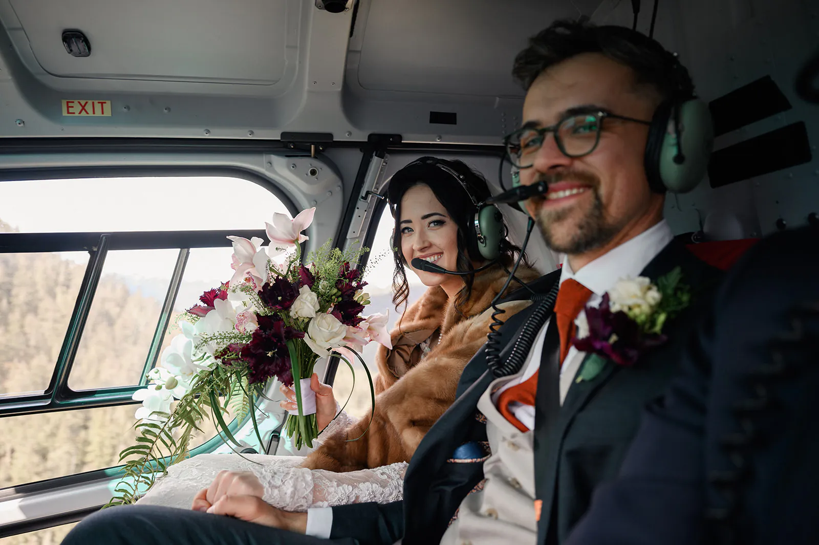 A bride and groom wearing headsets sit inside a helicopter, holding bouquets and smiling, dressed in wedding attire.