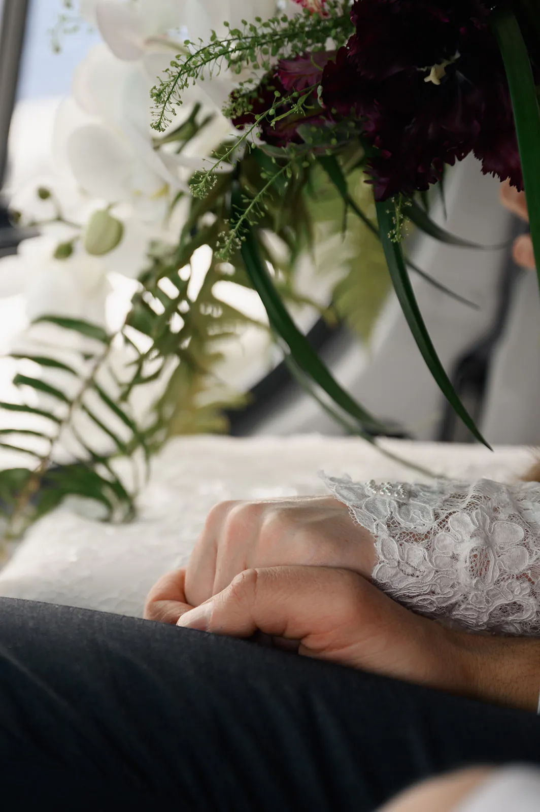 A close-up of two people holding hands, one wearing white lace, with a bouquet of green leaves and white flowers nearby.