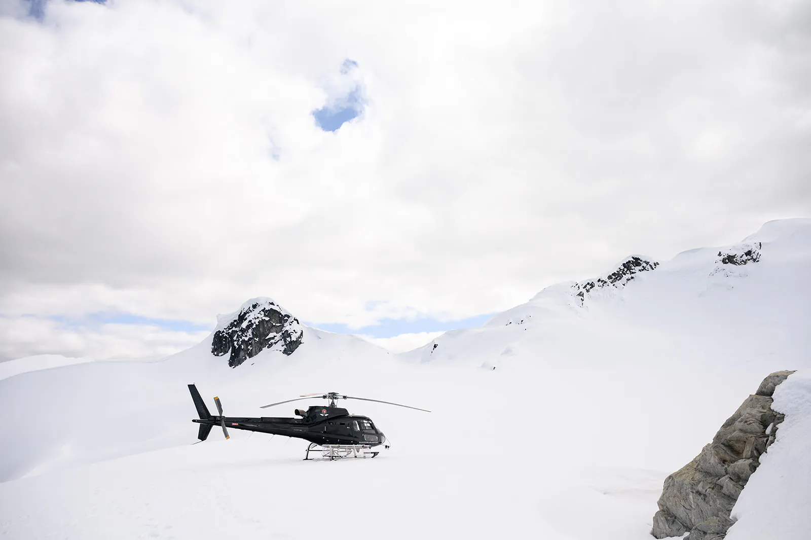 A black helicopter is parked on a snowy mountain landscape under a cloudy sky, with snow-covered peaks in the background.
