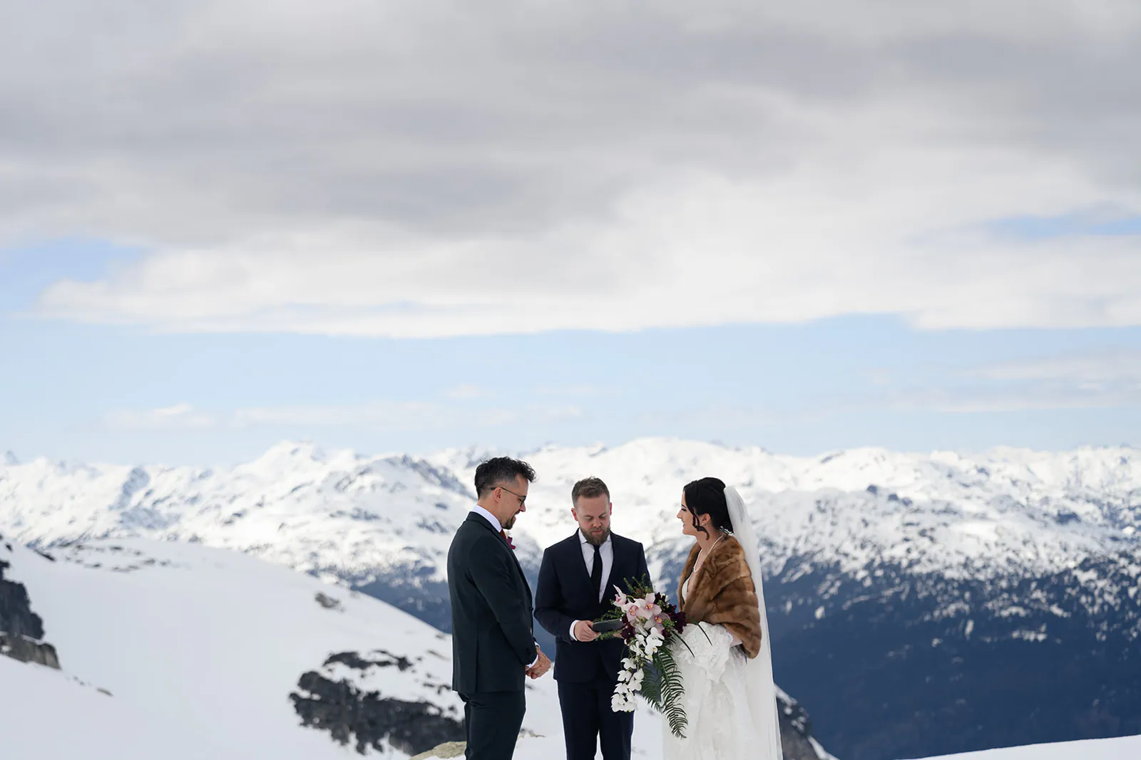 Three people stand outdoors on a snowy mountain during a wedding ceremony, with the officiant between the bride and groom. Snow-covered peaks and a cloudy sky are in the background.