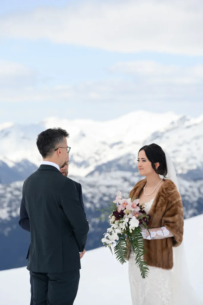 A bride and groom stand facing each other during an outdoor wedding ceremony on a snowy mountain, with the bride holding a bouquet and wearing a fur shawl.