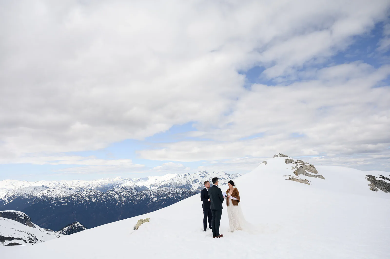 A couple stands with an officiant on a snowy mountain peak, surrounded by snow-covered landscapes and partly cloudy skies.