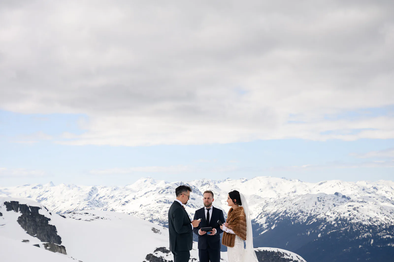 Three people stand together on a snowy mountain with a cloudy sky; one officiates while the other two appear to be exchanging wedding vows.