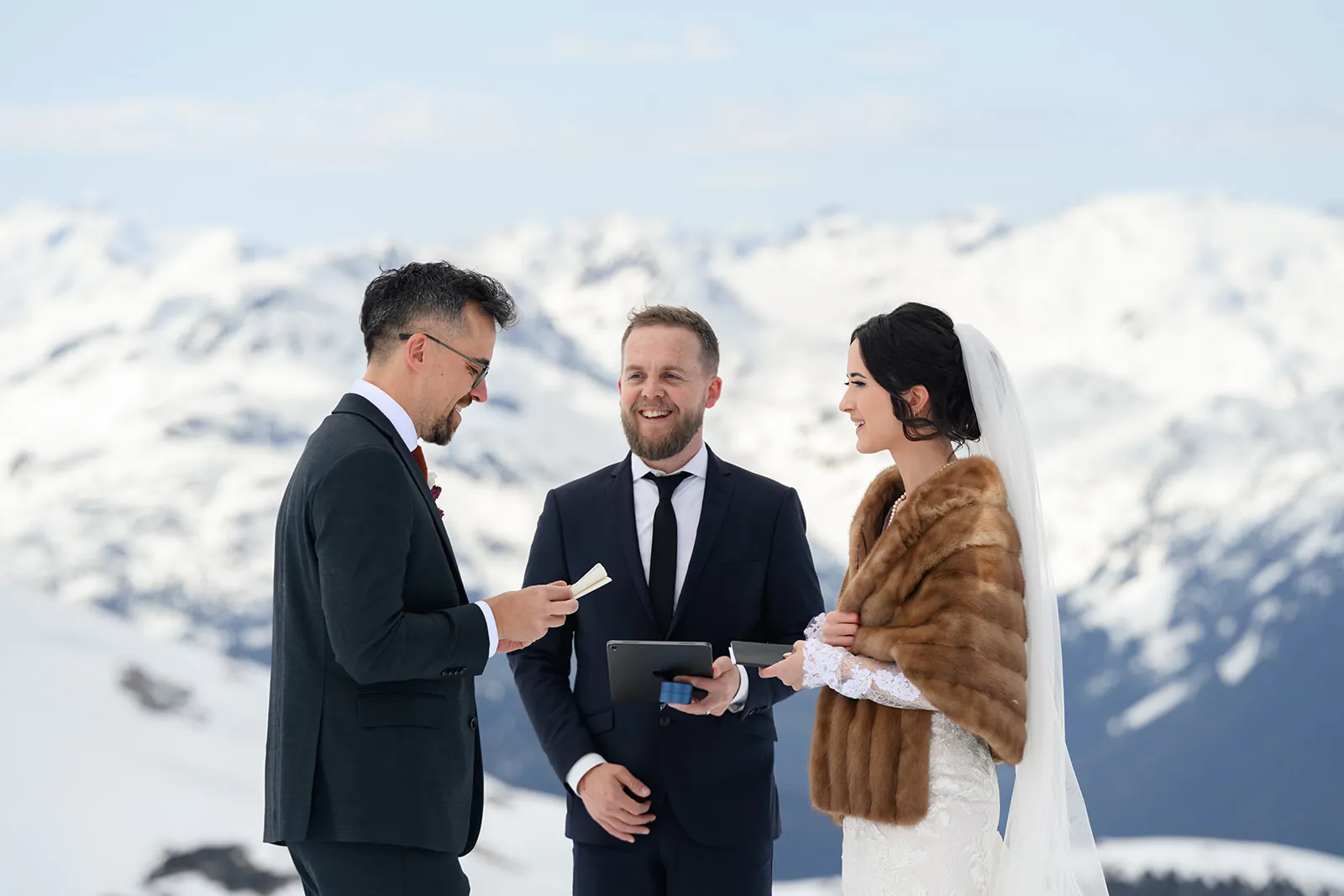 A couple exchanges vows during an outdoor wedding ceremony with an officiant, set against a snowy mountain backdrop.