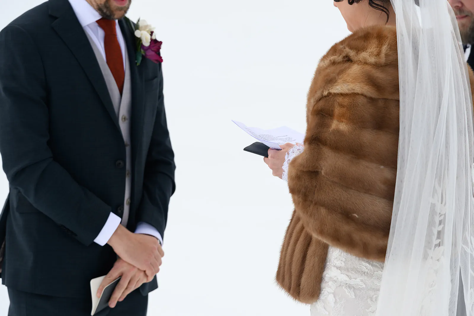 A bride in a fur shawl and veil reads from a piece of paper while standing next to a groom in a suit holding a notebook.