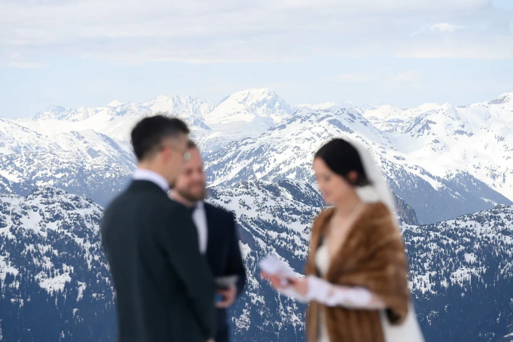 A bride and groom stand with an officiant in front of snow-covered mountains, likely during a wedding ceremony.