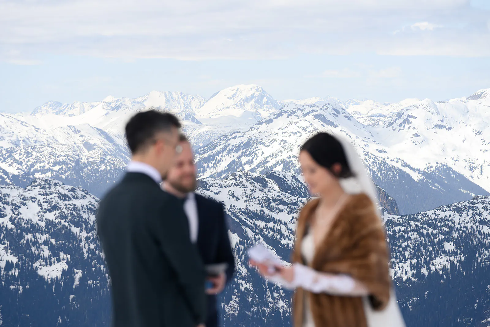 A bride and groom stand with an officiant in front of snow-covered mountains, likely during a wedding ceremony.