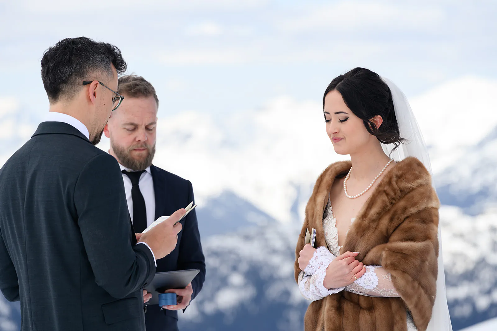 A bride and groom stand with an officiant during an outdoor wedding ceremony in a snowy mountain setting. The bride wears a fur stole and the groom reads from a phone.