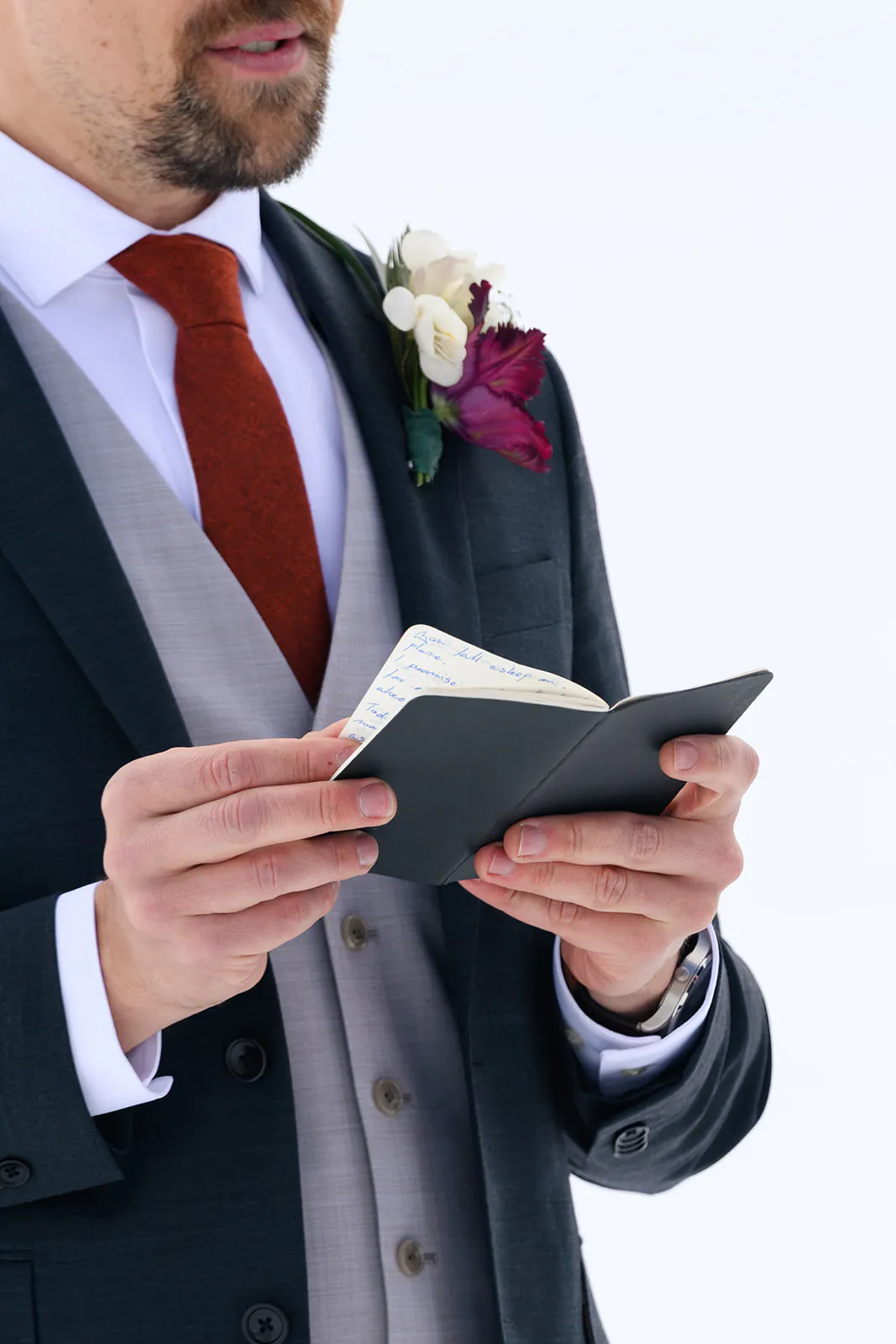 A man in a suit with a red tie and boutonniere holds an open notebook, reading from it.