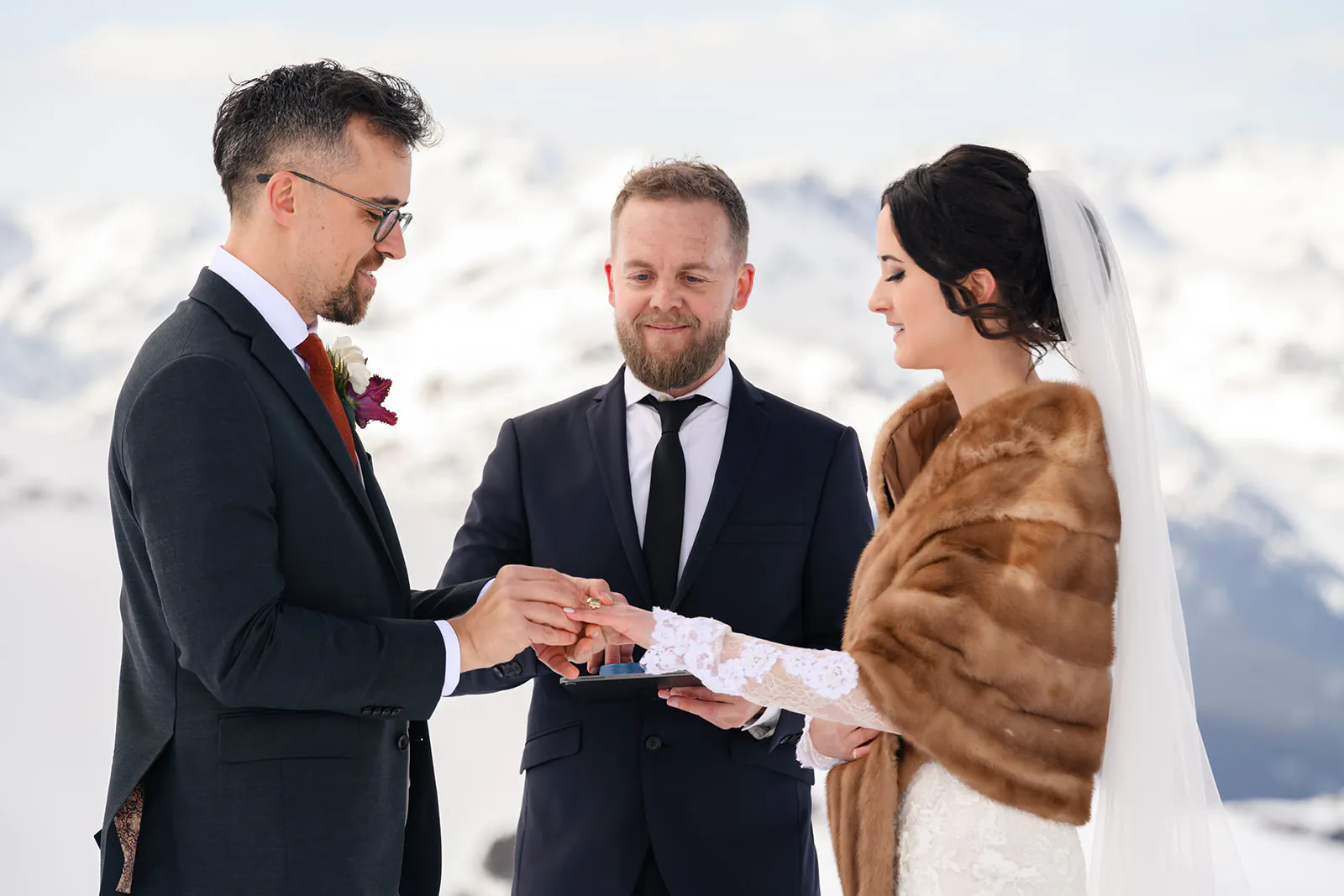 A bride and groom exchange rings during an outdoor wedding ceremony in a snowy mountain setting, with an officiant standing between them.