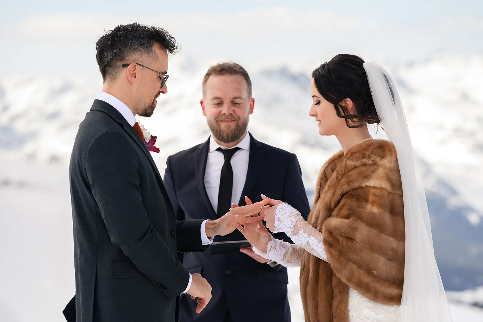 A bride and groom exchange rings during an outdoor wedding ceremony in a snowy mountain setting, officiant standing behind them.