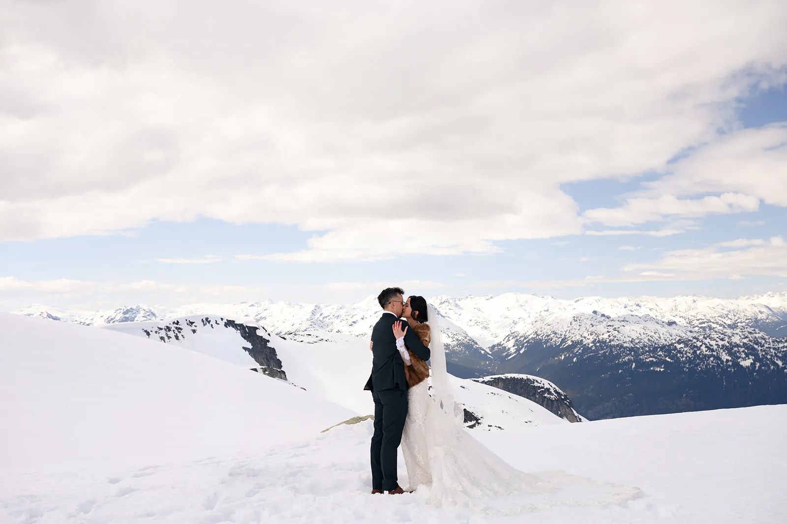 A bride and groom kiss on a snow-covered mountain with distant peaks and a cloudy sky in the background.