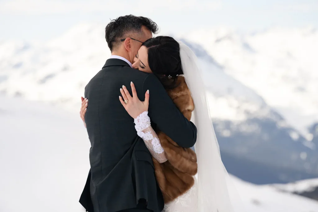 A bride and groom embrace outdoors in snowy mountains; the bride wears a veil and fur stole, and the groom is in a dark suit.