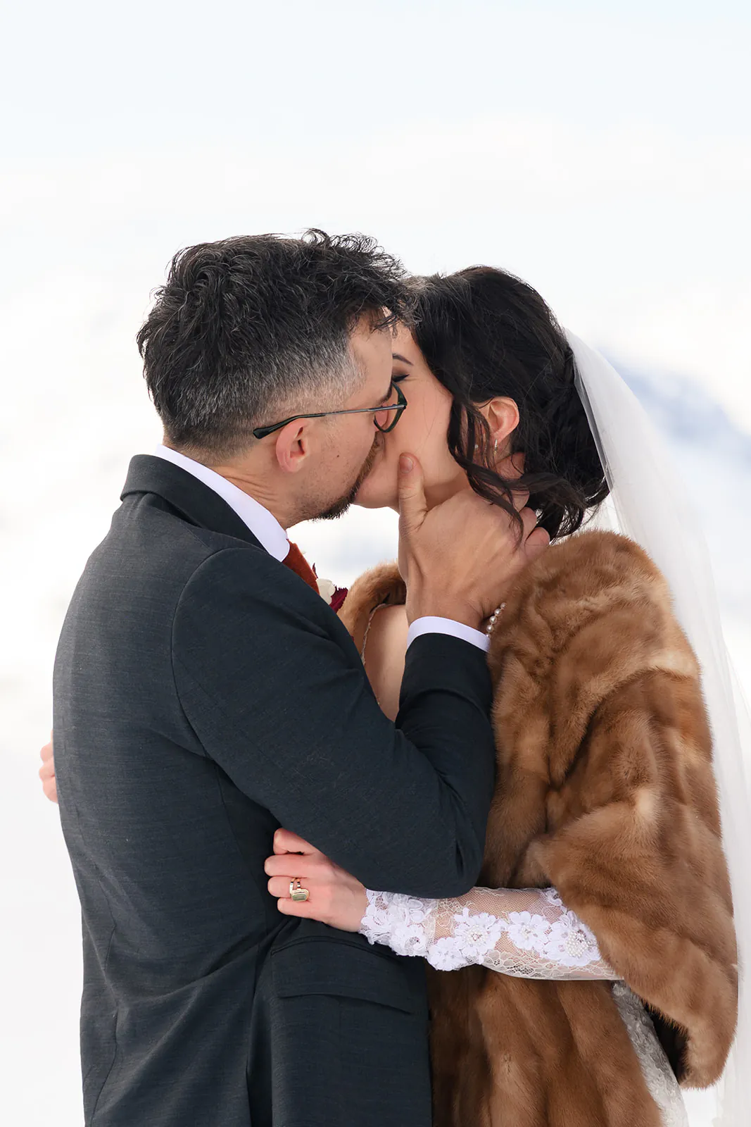 A bride and groom kiss outdoors; the bride wears a lace dress, brown fur shawl, and veil, while the groom wears a dark suit and glasses.