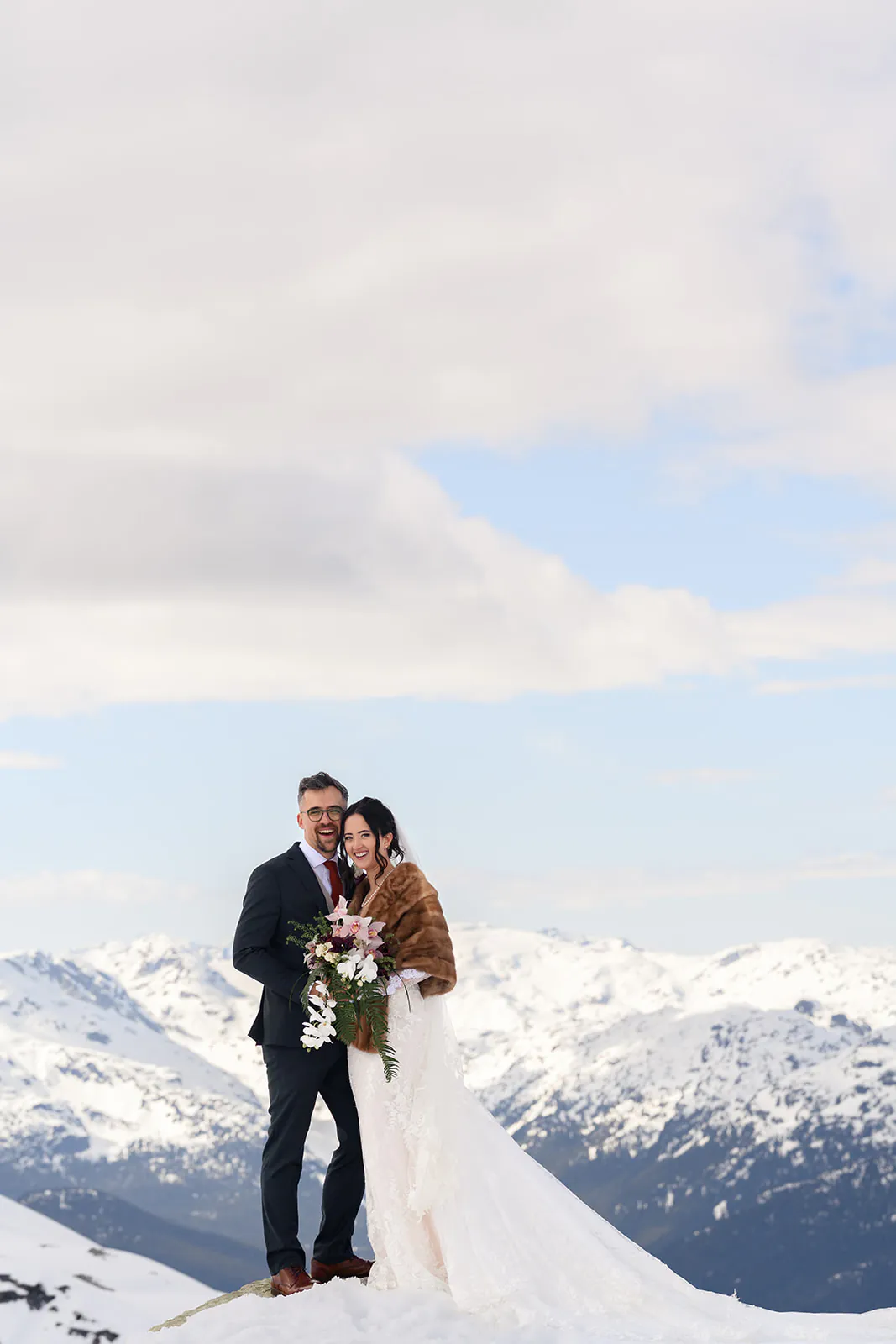 A bride and groom stand together on snowy ground, holding a floral bouquet, with snow-covered mountains and a cloudy sky in the background.
