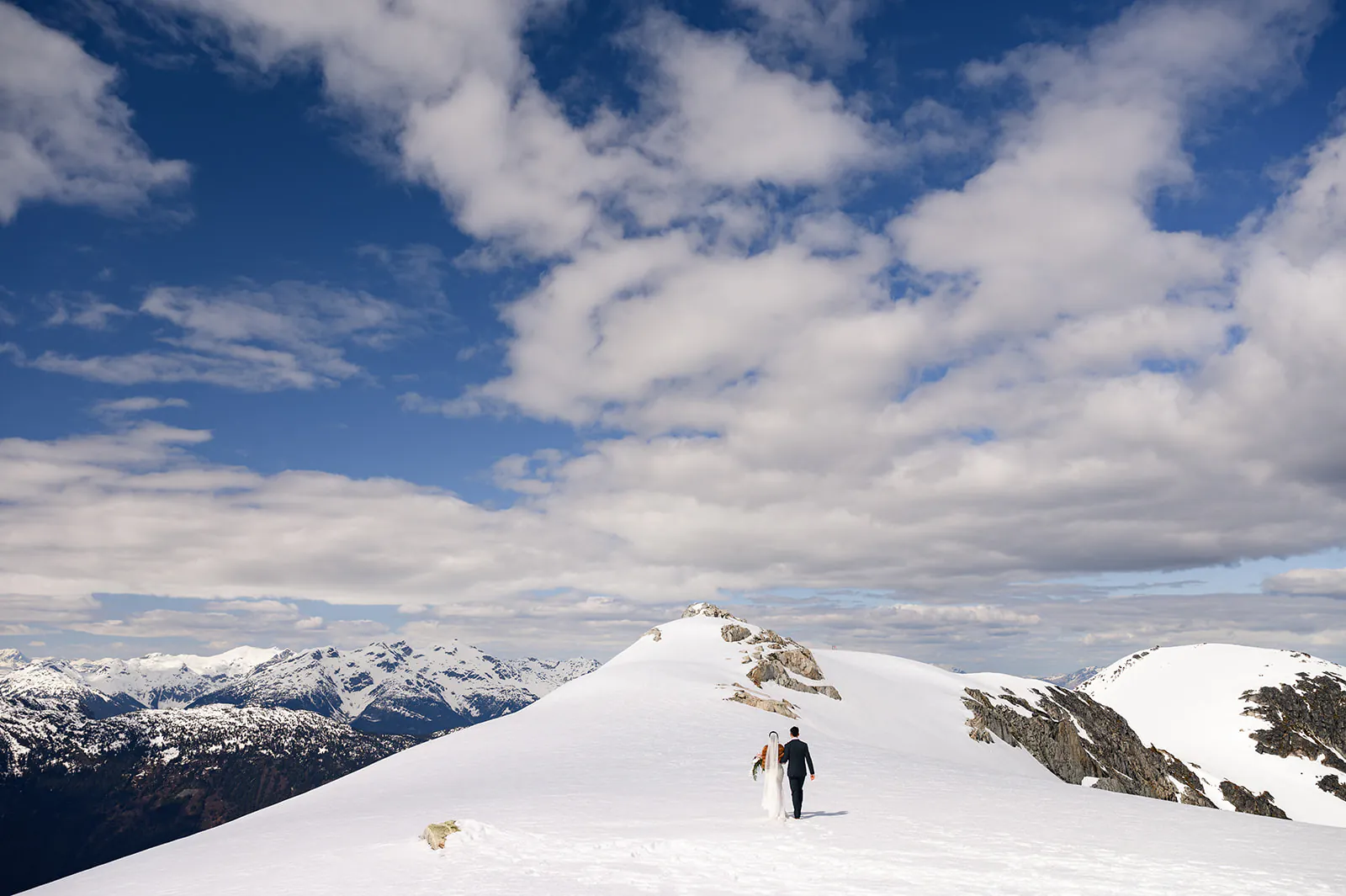 A couple dressed in wedding attire walks on a snow-covered mountain ridge under a partly cloudy sky with distant mountain ranges visible.