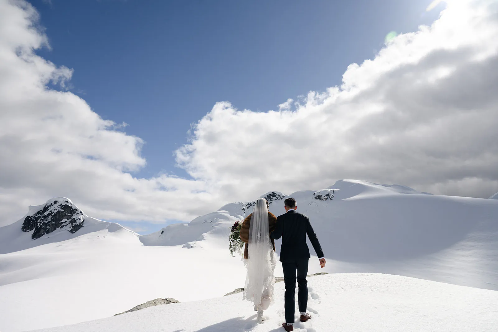 A couple dressed in formal wedding attire walks together on a snowy mountain landscape under a partly cloudy sky.