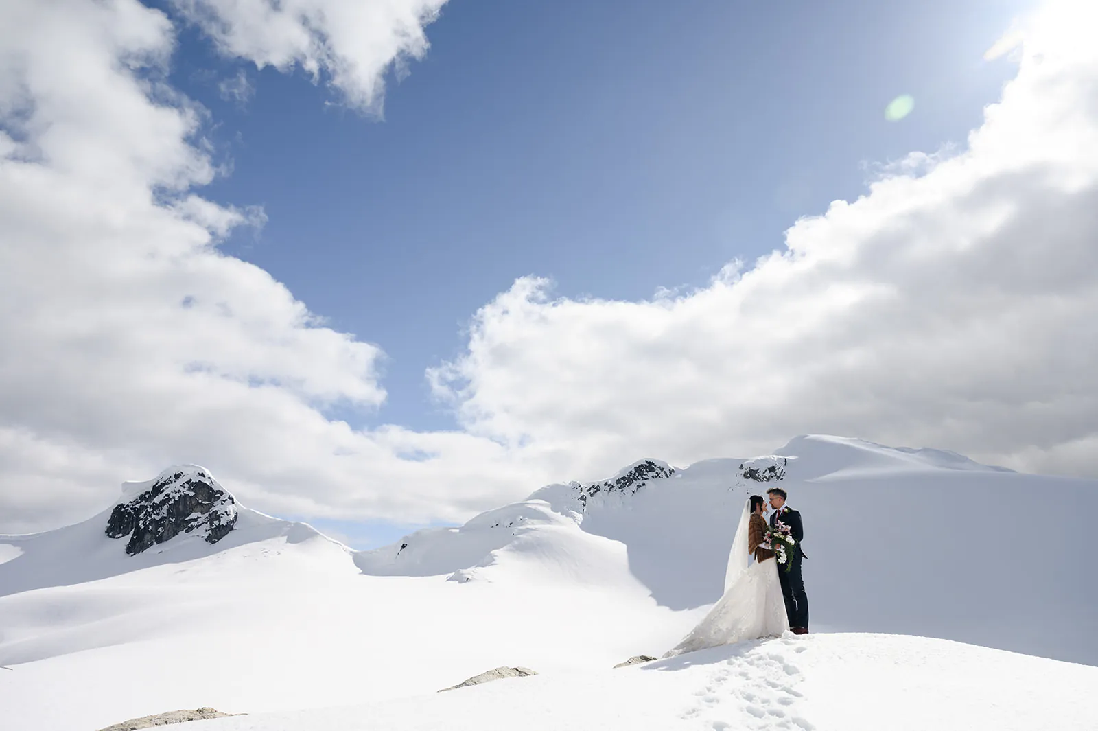 Bride and groom stand together on a snowy mountain peak under a partly cloudy sky with footprints leading up to them.