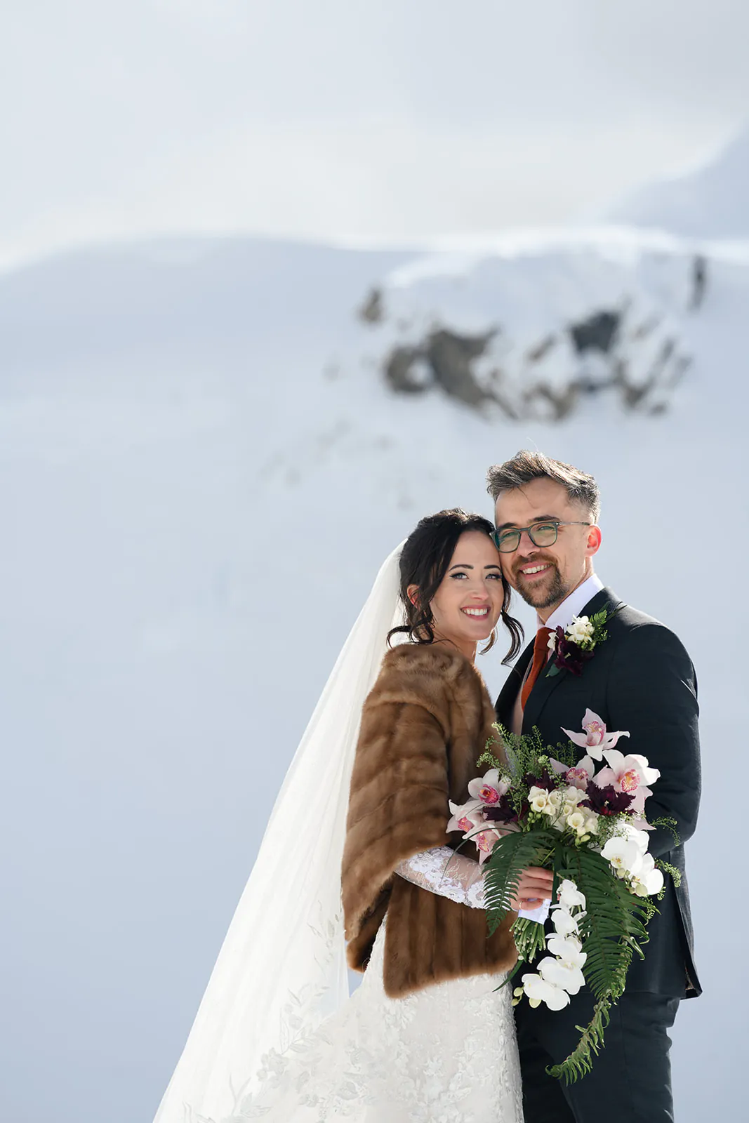 A bride and groom stand close together, smiling, with snow-covered mountains in the background. The bride wears a fur shawl and veil, while the groom holds a bouquet.