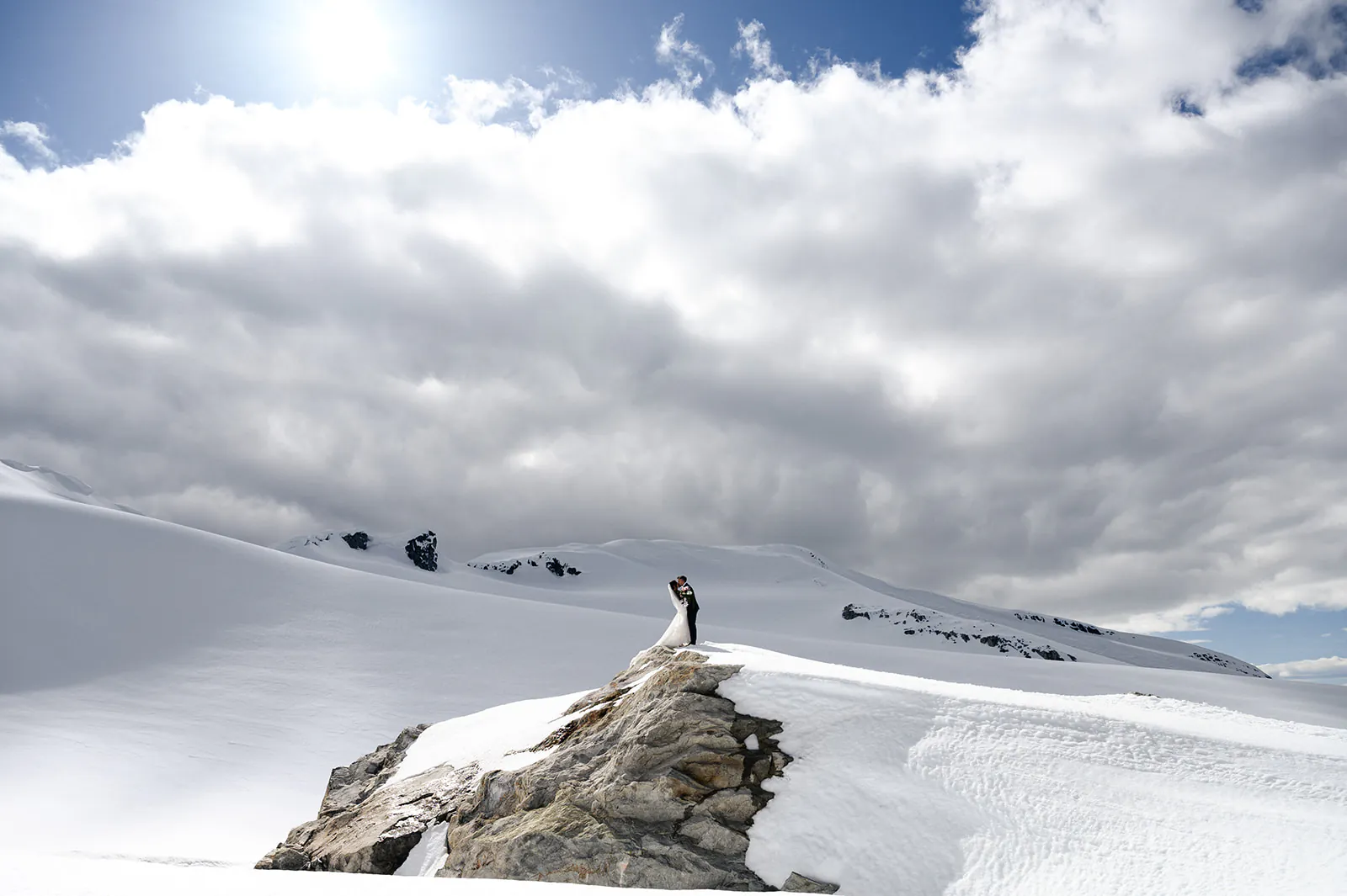 A couple stands and embraces on a rocky outcrop surrounded by snow-covered mountains under a partly cloudy sky.