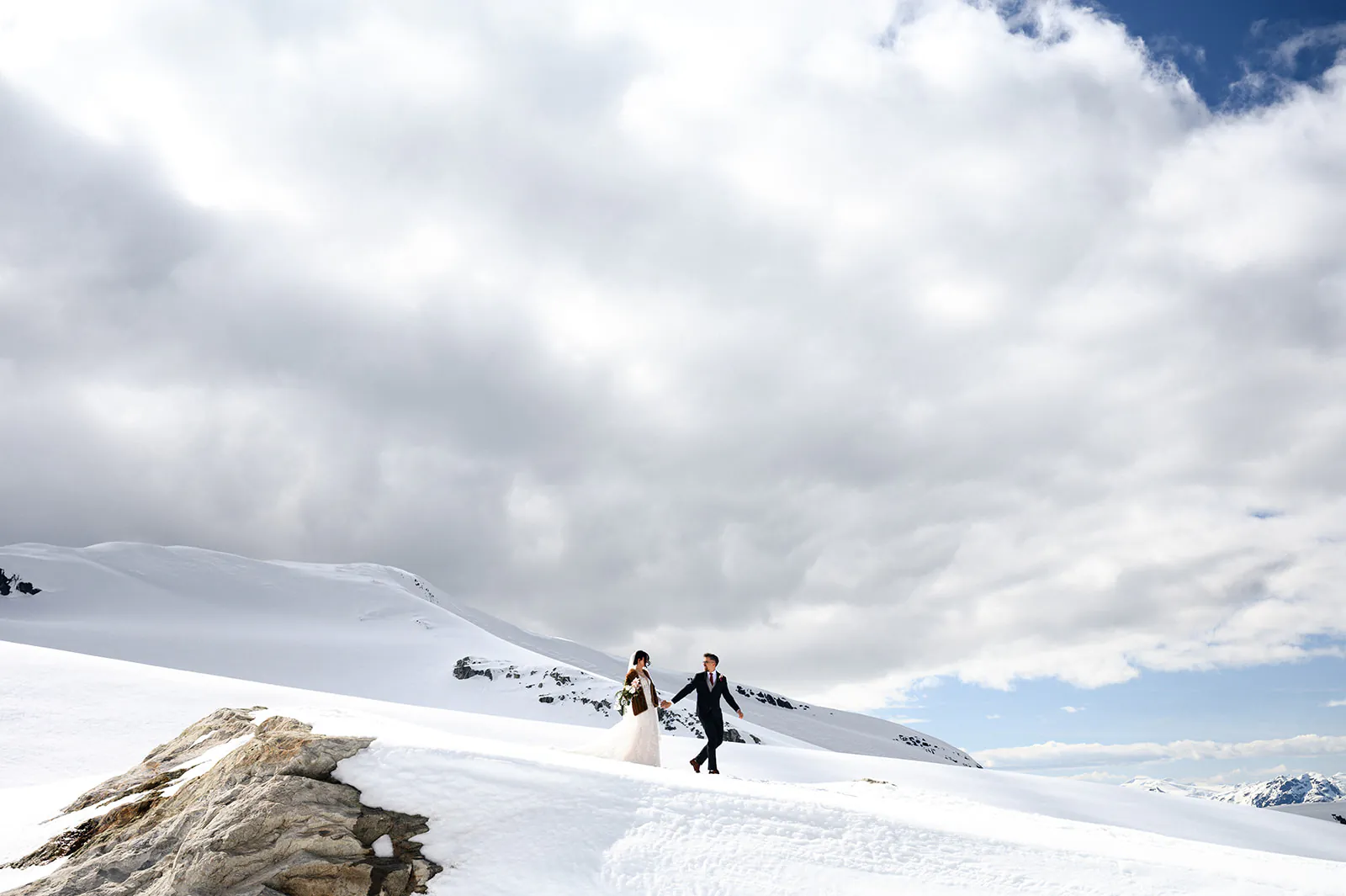 A bride and groom walk hand in hand across a snowy mountain landscape under a cloudy sky.