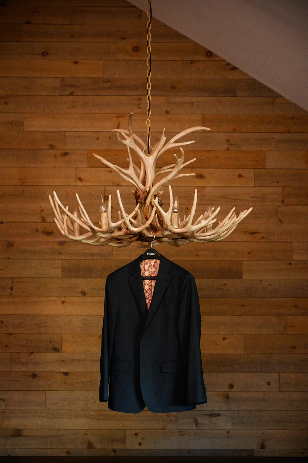 A dark suit jacket on a hanger is suspended from an antler chandelier against a wooden paneled wall.