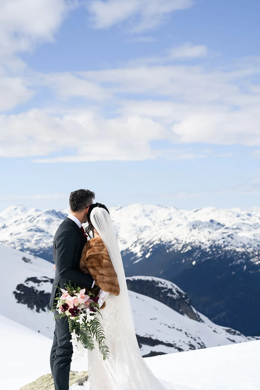 A bride and groom stand together on a snowy mountain, facing away and looking at the distant snow-covered peaks under a partly cloudy sky.