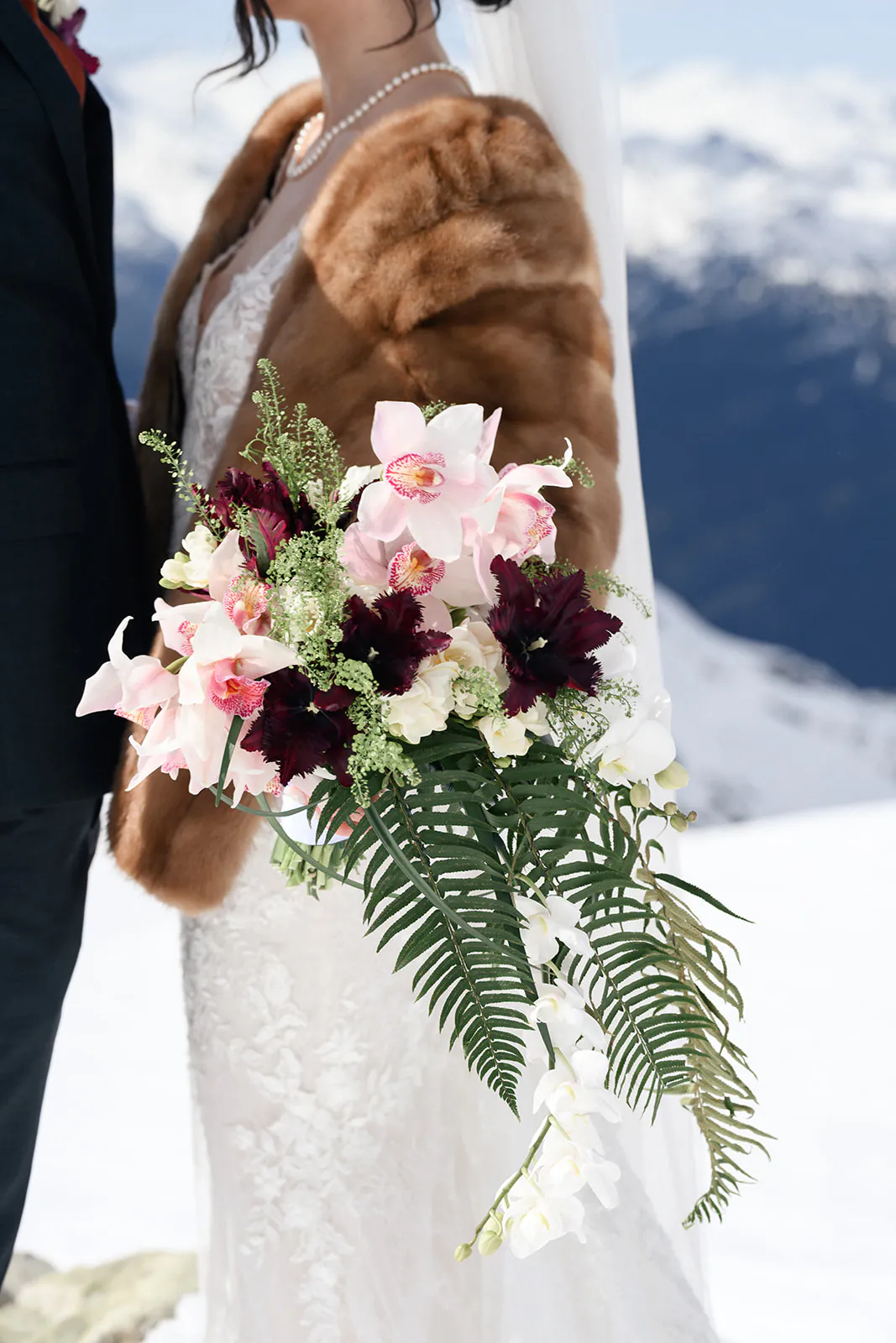 A bride in a fur shawl holds a cascading bouquet of pink, white, and dark burgundy flowers with greenery, standing outdoors in a snowy mountain setting.
