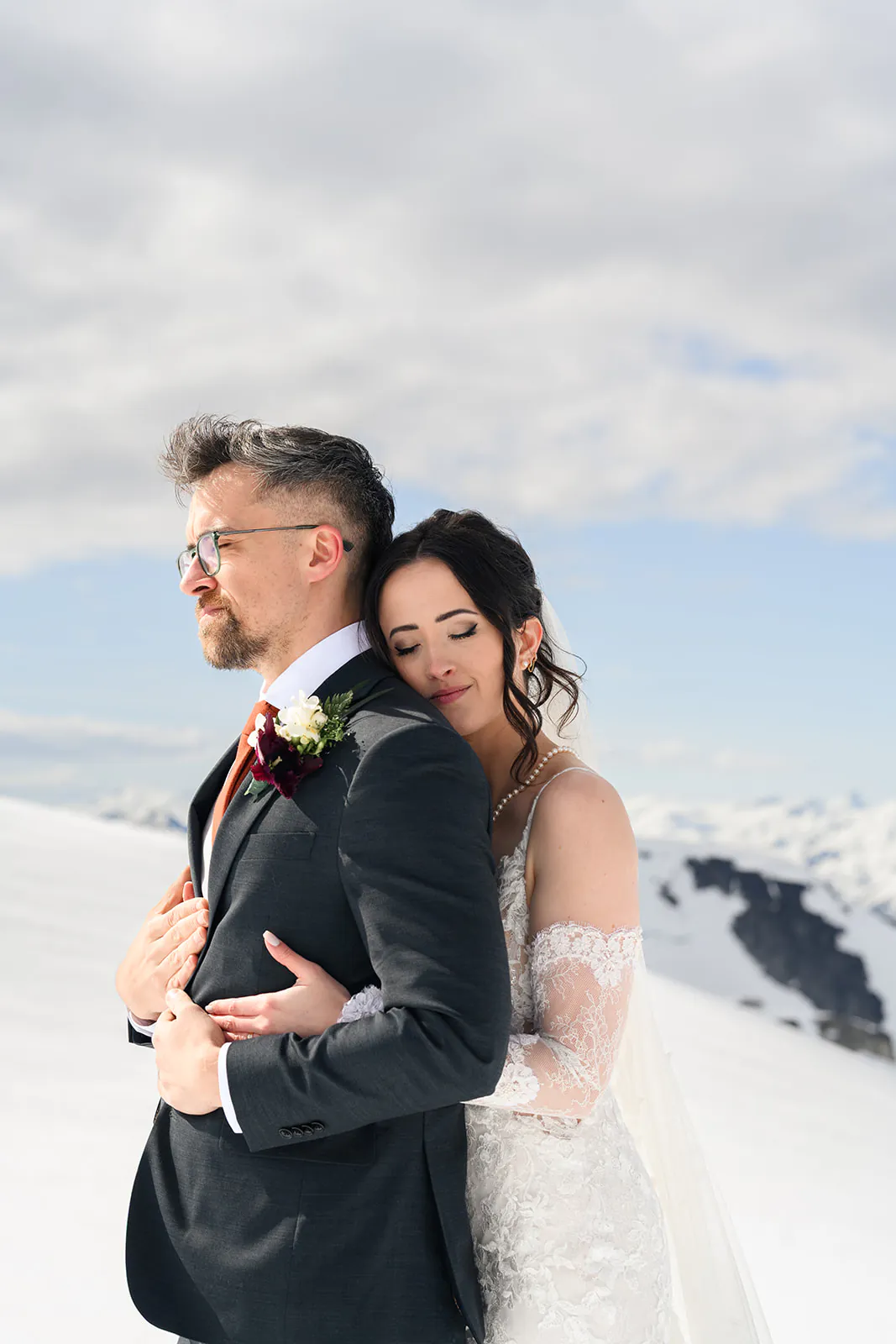 A bride in a white lace dress hugs a groom in a dark suit from behind; they stand on snow with mountains and a cloudy sky in the background.