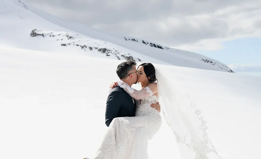 A bride and groom embrace and kiss in a snowy, mountainous landscape under a cloudy sky.