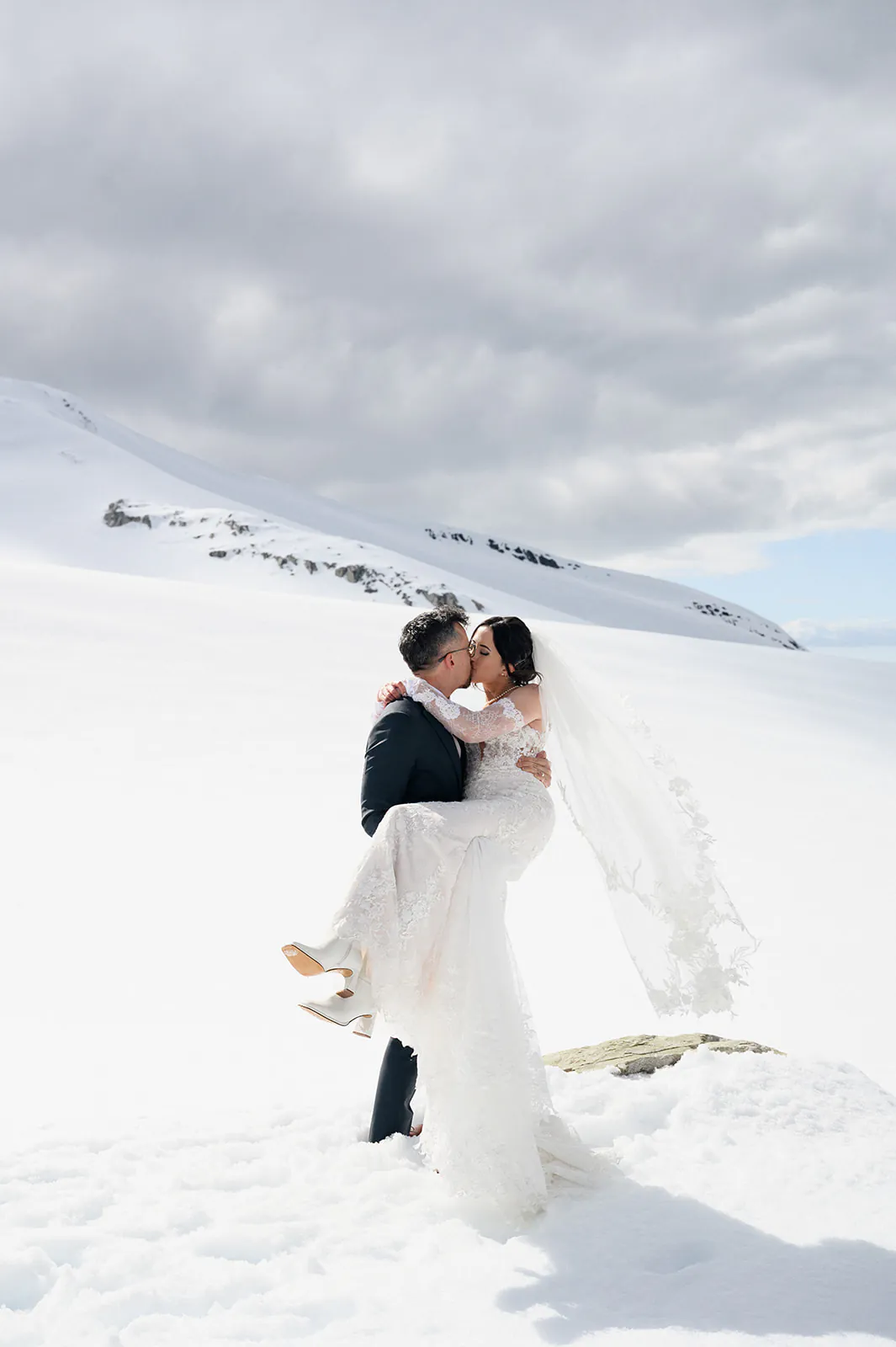 A bride and groom embrace and kiss in a snowy, mountainous landscape under a cloudy sky.