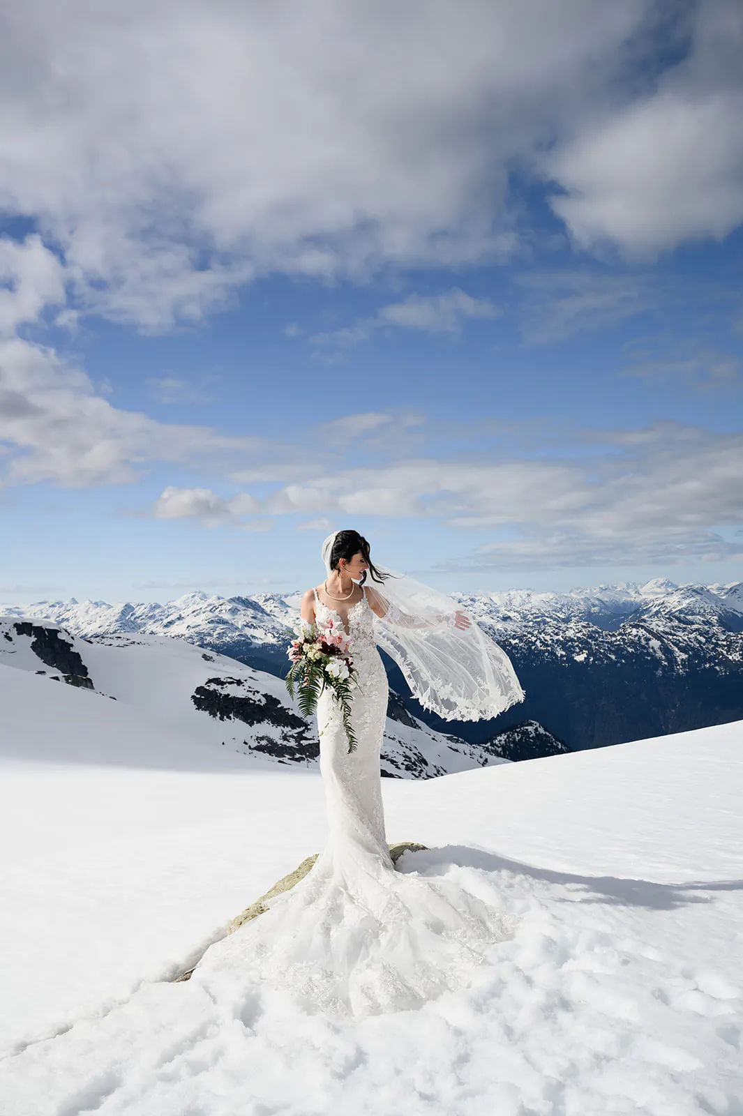 A bride in a white wedding dress stands on snow-covered ground, holding a bouquet, with mountains and a blue sky in the background.