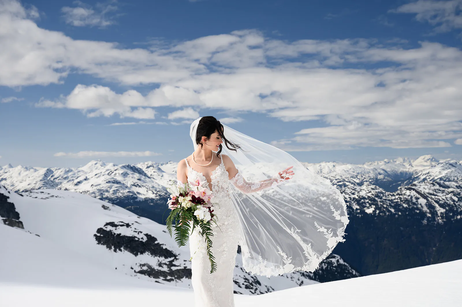 A bride in a white dress and veil holds a bouquet while standing on a snowy mountain with a scenic background of snow-covered peaks under a partly cloudy sky.