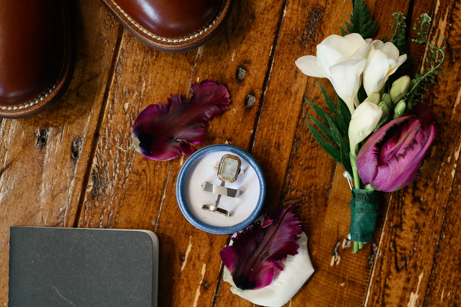 Cufflinks in a box, a boutonniere, flower petals, and brown shoes arranged on a wooden surface.