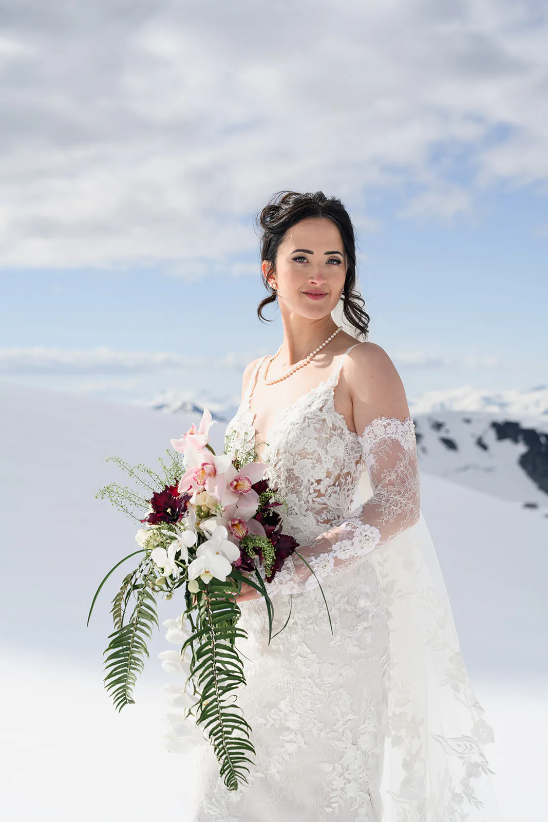 A bride in a lace wedding dress holding a bouquet of flowers stands outdoors on a snowy mountain under a cloudy sky.