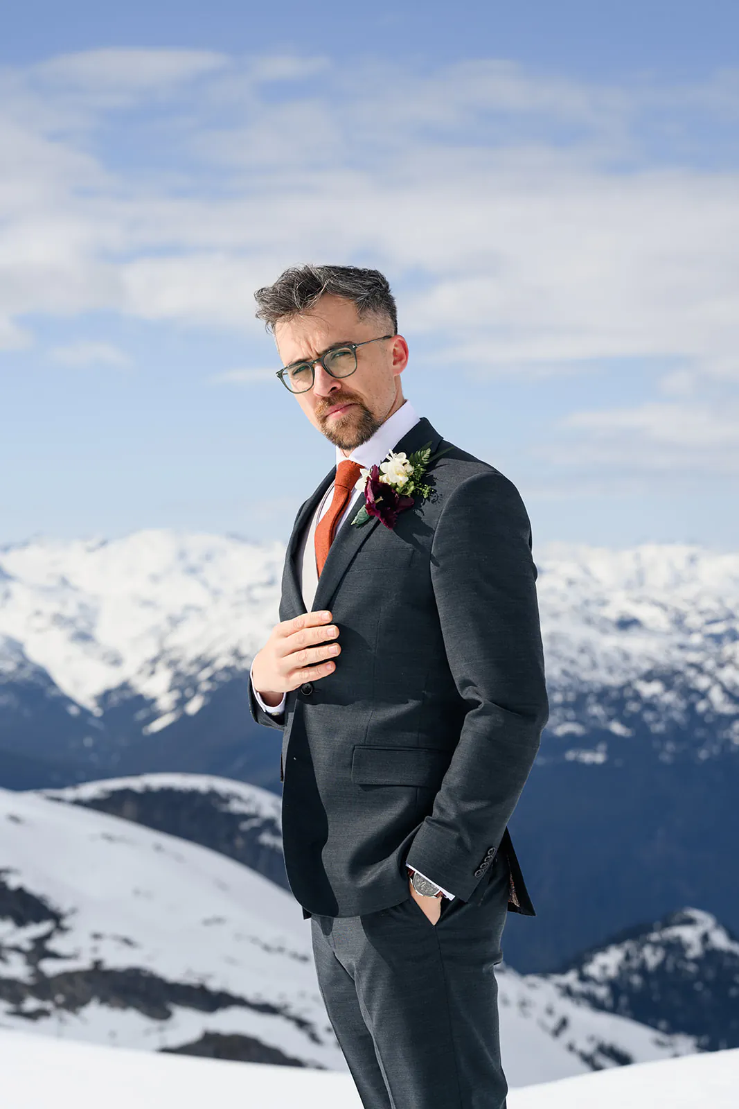 A man in a dark suit with a boutonnière stands on a snowy mountain, with snow-covered peaks and a blue sky in the background.