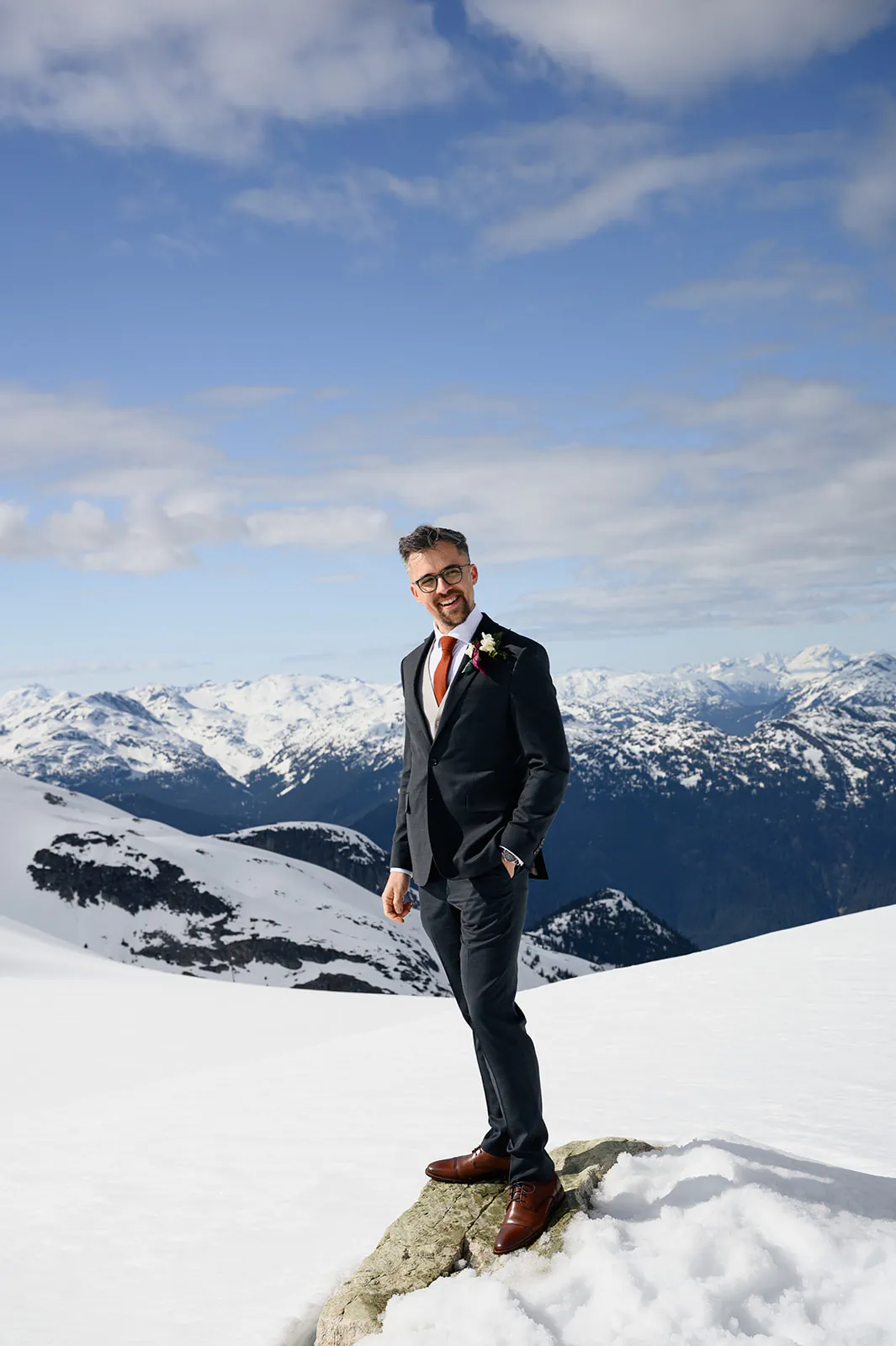 A man in a suit with a boutonnière stands on a rock in a snowy mountain landscape under a partly cloudy sky.