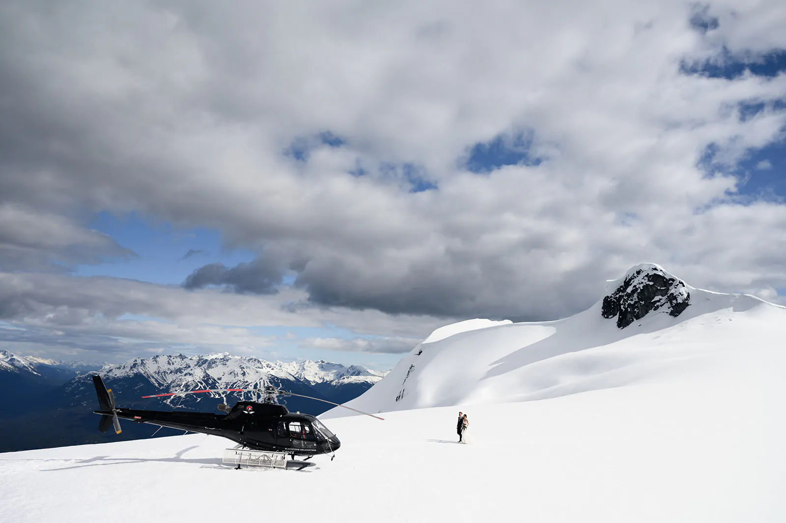 A black helicopter is parked on a snowy mountain slope under a partly cloudy sky, with a person standing nearby and snow-covered peaks in the background.