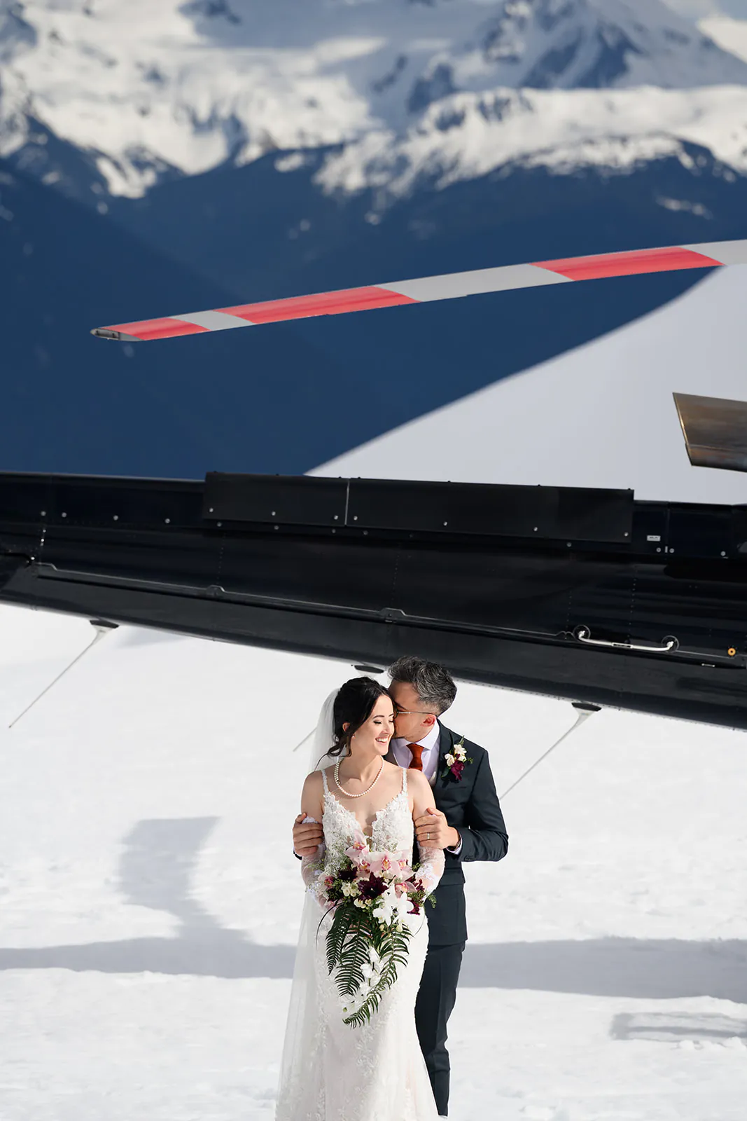 A bride and groom stand together in snow near a helicopter, with mountains in the background. The bride holds a bouquet of pink flowers and greenery.