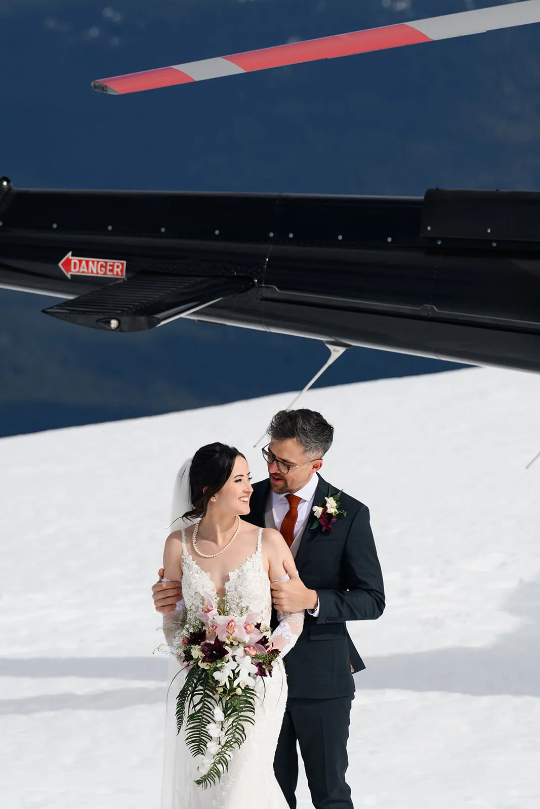 A bride and groom stand together on snow near a black helicopter, with the groom holding the bride from behind and both smiling.