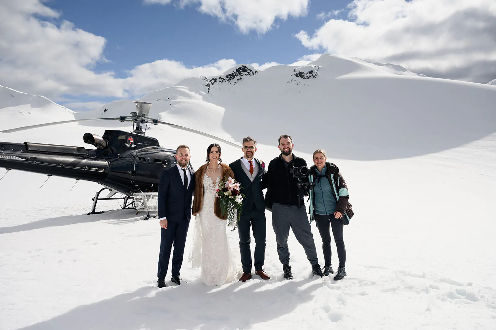 Five people stand on snow in front of a black helicopter and snow-covered mountains, with one person in a wedding dress holding a bouquet.