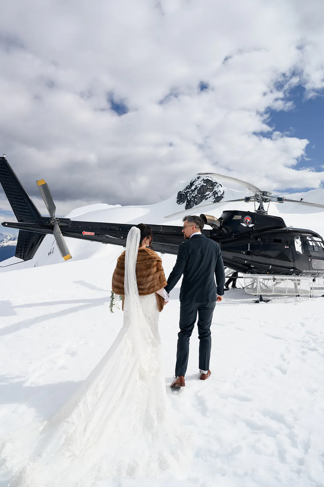 A bride and groom stand on snow next to a black helicopter, holding hands and facing away, with mountains and clouds in the background.